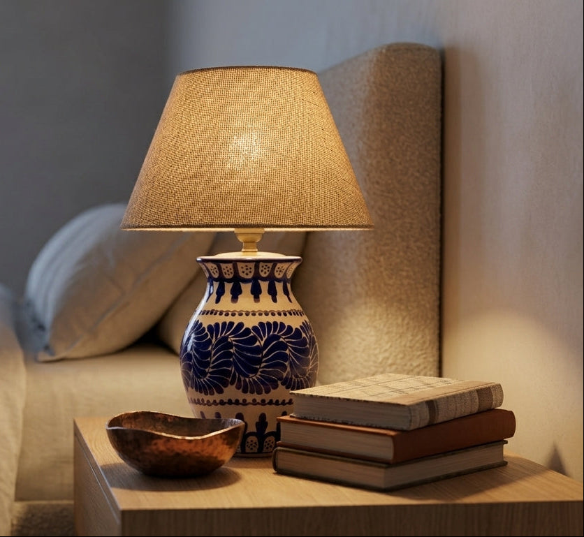 A lit table lamp with a beige fabric shade and a blue-and-white patterned ceramic base sits on a wooden bedside table next to stacked books and a bowl in a softly lit bedroom.
