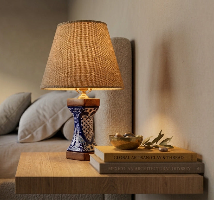 A warm lit table lamp with a beige fabric drum shade and a blue-and-white patterned ceramic base sits on a wooden nightstand beside a bed in a softly lit bedroom.